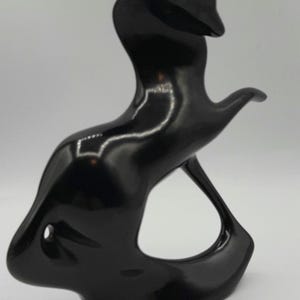 May include: A glossy black abstract sculpture with a smooth, curved design. The sculpture has a flowing form with a rounded base, a central opening, and an upward-reaching element. The surface reflects light, highlighting its shape.