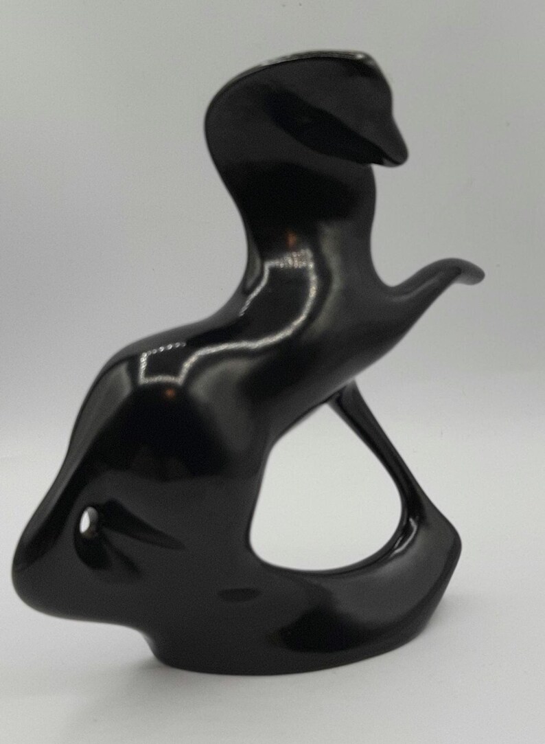 May include: A glossy black abstract sculpture with a smooth, curved design. The sculpture has a flowing form with a rounded base, a central opening, and an upward-reaching element. The surface reflects light, highlighting its shape.