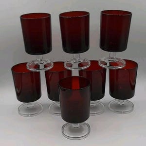 May include: A set of seven matching stemmed glasses featuring deep red, translucent bowls and clear glass bases. Each glass has a cylindrical shape with a smooth rim, supported by a short, sturdy clear stem and a circular footed base.