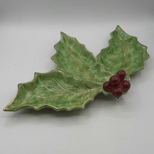 Puede incluir: Un plato de cerámica con forma de hoja de acebo, con acabado verde brillante y ribete dorado. El plato presenta tres secciones de hojas y un racimo de bayas rojas. Ideal para servir dulces navideños o como adorno decorativo.