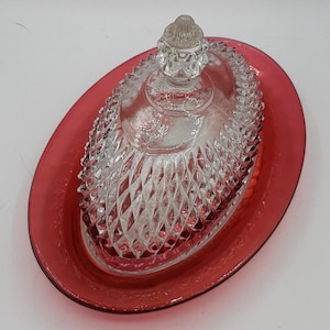 Può includere: Una burriera ovale in vetro rosso rubino con coperchio in vetro trasparente tagliato a diamante. Il coperchio ha un pomello decorativo e poggia su una base rossa abbinata. Il piatto è progettato per servire il burro.