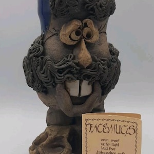 May include: A brown ceramic mug shaped like a face with a textured mustache, eyebrows, and two prominent front teeth. The mug has a blue rim and is accompanied by a small tan card that reads FACEMUGS, oven proof, water tight, lead free, dishwasher safe, by McGillivray Crafts.