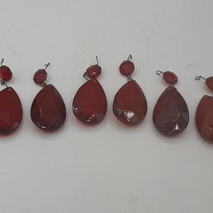 Puede incluir: Seis cristales de araña de lágrimas rojas con superficies facetadas. Cada cristal tiene una cuenta roja redonda más pequeña encima, conectada por un bucle de alambre. Los cristales están dispuestos en una fila sobre un fondo blanco.