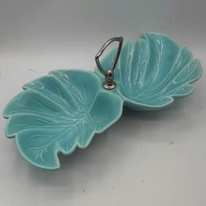 May include: A turquoise ceramic leaf-shaped serving dish with a silver handle. The dish is divided into two sections, each with detailed leaf vein patterns. The handle is arched and attached to a central base. The dish is likely vintage.