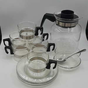 May include: A clear glass coffee set featuring a carafe, six cups with black handles, saucers, and a small bowl with a spoon. The cups have silver metal accents. Ideal for serving coffee or tea.