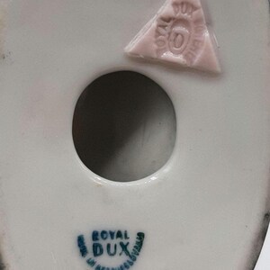 May include: Close-up of the underside of a white porcelain figurine with a dark gray rim. Features a circular hole and a pink triangular stamp with text. The base has a blue and green "ROYAL DUX" stamp.