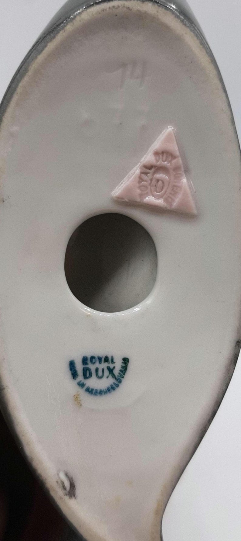 May include: Close-up of the underside of a white porcelain figurine with a dark gray rim. Features a circular hole and a pink triangular stamp with text. The base has a blue and green "ROYAL DUX" stamp.
