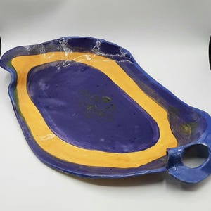 Può includere: Un vassoio da portata in ceramica fatto a mano con una forma organica unica. Il vassoio presenta un interno blu scuro, un bordo giallo vibrante e un manico blu. I bordi hanno una finitura strutturata e macchiettata, che aggiunge al suo fascino artigianale.