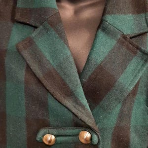 May include: Green and black checked blazer with gold buttons. The double-breasted jacket features a traditional notched lapel and a fitted silhouette. The material looks like a wool mix, ideal for colder climates. A vintage-style piece of clothing.