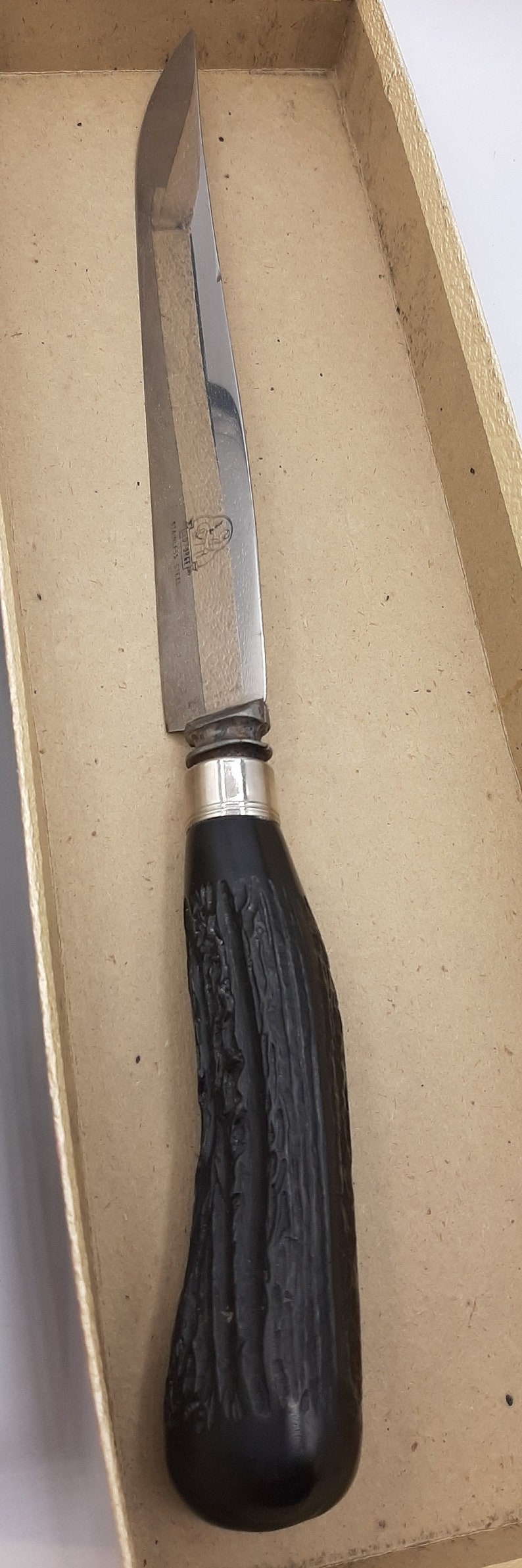 Puede incluir: Un cuchillo de caza vintage con una hoja de acero inoxidable y un mango oscuro y texturizado. El cuchillo est&aacute; en una caja de cart&oacute;n. La hoja tiene las palabras "STAINLESS STEEL" y un logotipo.