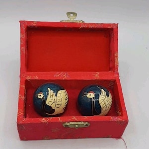 Peut inclure: Deux boules métalliques sphériques avec une finition émaillée bleu foncé, chacune présentant un motif de grue blanche et noire avec une couronne rouge. La paire est logée dans une boîte rectangulaire recouverte de tissu rouge avec un fermoir en métal.