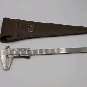 Puede incluir: Un calibrador de metal plateado con una escala de medición, junto a un estuche de cuero marrón. El calibrador tiene la marca "MEBA". El estuche está cerrado con una correa.