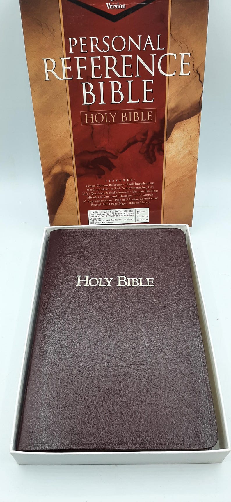 May include: A brown leather-bound Holy Bible with gold lettering, displayed in a white box. The cover reads "HOLY BIBLE" in gold. The box behind it displays "PERSONAL REFERENCE BIBLE" and "HOLY BIBLE".