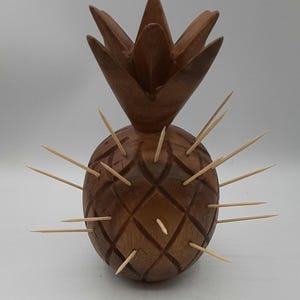 Könnte beinhalten: Ein hölzerner Zahnstocherhalter in Ananasform. Die braune Holzananas hat ein geschnitztes Rautenmuster und Holz-Zahnstocher, die um den Körper herum eingesetzt sind. Die Oberseite der Ananas ist eine detaillierte Krone.