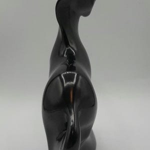 May include: A glossy black ceramic cat sculpture. The cat is depicted in a stylized, abstract form, with a curved neck and a curled tail. The sculpture has a smooth, reflective surface.