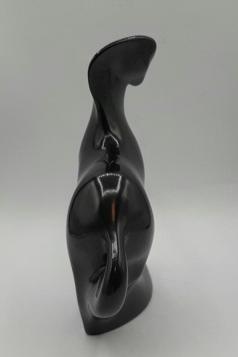 May include: A glossy black ceramic cat sculpture. The cat is depicted in a stylized, abstract form, with a curved neck and a curled tail. The sculpture has a smooth, reflective surface.