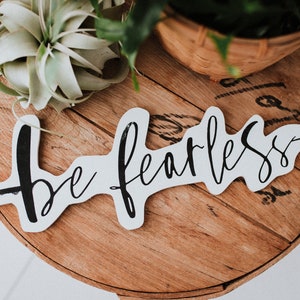 Puede incluir: Un letrero de madera blanco con caligrafía negra que dice "be fearless" con una flecha en cada extremo. El letrero está sobre una mesa de madera con una planta verde en una cesta tejida.