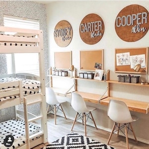 Puede incluir: Una litera blanca con ropa de cama con estampado blanco y negro. Tres sillas blancas con patas de madera est&aacute;n colocadas frente a un escritorio con una tapa de madera y soportes de metal blanco. Tres tableros de corcho con nombres escritos en ellos est&aacute;n en la pared detr&aacute;s del escritorio. Los nombres son "Sydney Blair", "Carter Blake" y "Cooper Allen".