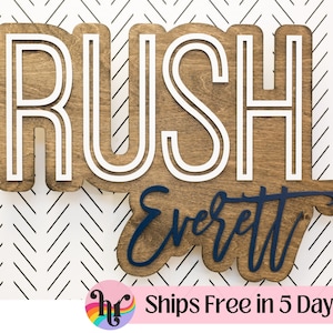 May include: A wooden wall sign featuring the word RUSH in large, white, block-style letters layered over a dark wood-stained backing. Below, the name Everett is written in a dark blue, cursive script font, also layered onto the wood base.