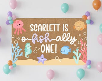Può includere: Un cartello marrone con la scritta "Scarlett is o-fish-ally ONE!" è circondato da palloncini colorati. Il cartello presenta creature marine come una medusa, un cavalluccio marino e un pesce. Palloncini nei toni del rosa, arancione e verde acqua sono disposti intorno.