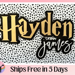 May include: A black and gold wooden sign with the name "Hayden James" in a decorative font. The sign is on a white background with black polka dots.