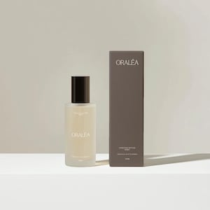 Op de afbeelding: Een fles van matglas met ORALÉA Hydrating Setting Spray en een zwarte dop, naast een bijpassende rechthoekige doos. De doos en de fles hebben het ORALÉA-logo. De doos vermeldt ook de ingrediënten en het volume van het product.