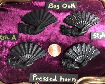 May include: Four black fan-shaped decorative objects, each held by a sculpted hand, are displayed on a purple velvet surface. The fans are labeled "Style A" and "Style B", with the text "Bog Oak" and "Pressed horn" also present.