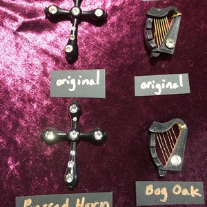 May include: A display of two types of jewelry. One is a black cross with clear crystal accents, labeled "Pressed Horn." The other is a black harp with gold strings, labeled "Bog Oak." Both are displayed on a burgundy velvet surface.