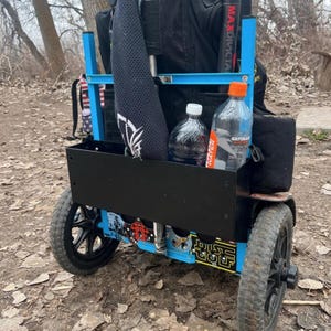 May include: A blue and black disc golf cart with black wheels. The cart has a black storage bin and holds water bottles and a black sleeve. The cart has the words "DISC GOLF" on the side.