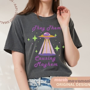 May include: A dark grey t-shirt with a graphic of a purple and yellow UFO with the text "They Them Causing Mayhem" in purple and yellow lettering.