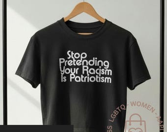 May include: Black t-shirt with the text "Stop Pretending Your Racism Is Patriotism" in white. The shirt is hanging on a wooden hanger. The words "march for the movement original design" are also visible.