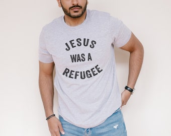 May include: A person wearing a grey t-shirt with the text "JESUS WAS A REFUGEE" printed on it in black.