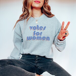 May include: A gray sweatshirt with the text "votes for women" in blue and orange letters. The person wearing the sweatshirt is sitting on a chair and making a peace sign with their hand.