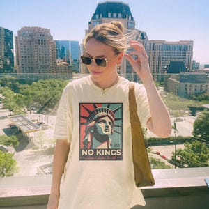 May include: Cream-colored t-shirt featuring a graphic of the Statue of Liberty with the text "NO KINGS" and "Liberty & Justice For All". The shirt is worn outdoors with a city backdrop.