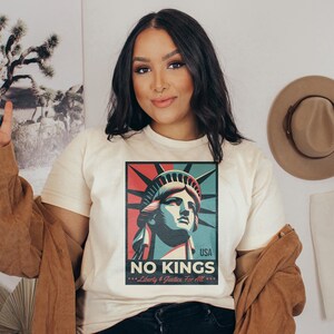 May include: Cream-colored t-shirt featuring a graphic of the Statue of Liberty in red, blue, and green, with the text "NO KINGS" and "Liberty & Justice For All." A brown jacket is draped over the shoulders.