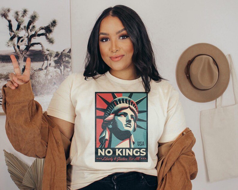 May include: Cream-colored t-shirt featuring a graphic of the Statue of Liberty in red, blue, and green, with the text "NO KINGS" and "Liberty & Justice For All." A brown jacket is draped over the shoulders.