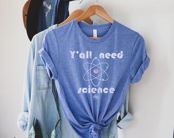May include: A blue t-shirt with the text "Y'all need science" and an atom graphic. The shirt is hanging on a wooden hanger with a denim shirt and a brown hat in the background. The shirt is made of a soft material.