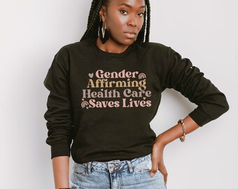 May include: A black sweatshirt with the text "Gender Affirming Health Care Saves Lives" in pink and gold lettering. The text is in a retro font style. The model is wearing blue jeans.