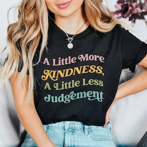 Può includere: Una maglietta nera con una grafica colorata che recita "A Little More Kindness, A Little Less Judgement".
