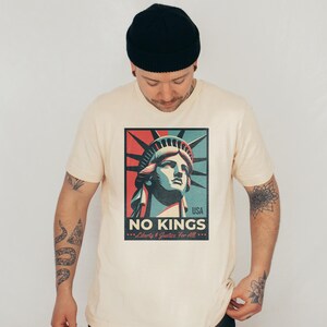 May include: Cream-colored t-shirt featuring a graphic of the Statue of Liberty with the text "NO KINGS" and "Liberty & Justice For All." The graphic uses red, blue, and green colors. The shirt is a crew neck.