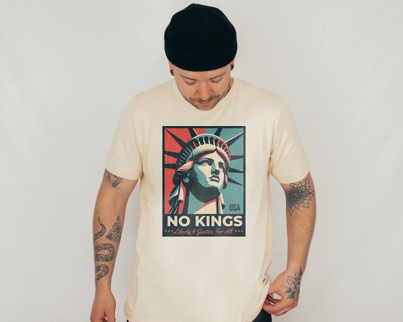 May include: Cream-colored t-shirt featuring a graphic of the Statue of Liberty with the text "NO KINGS" and "Liberty & Justice For All." The graphic uses red, blue, and green colors. The shirt is a crew neck.