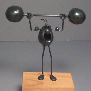 May include: A black metal sculpture of a cartoon character lifting weights. The character has a large head with a wide mouth and large eyes. The sculpture is mounted on a wooden base.