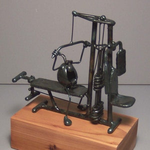 May include: A metal sculpture of a weightlifting machine with a figure sitting on the bench. The sculpture is painted in a dark metallic color and is mounted on a wooden base.