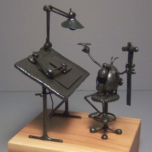 May include: A handcrafted metal sculpture of an artist at a drafting table. The sculpture features a desk with a lamp, a seated figure, and a wooden base. The artist figure is holding a pen and a tool.