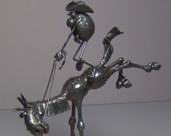 May include: A metal sculpture of a cowboy riding a bucking horse. The sculpture is made of wire and metal and is painted in a dark metallic color. The cowboy is wearing a hat and has his arms raised in the air. The horse is bucking and has its legs in the air. The sculpture is mounted on a wooden base.