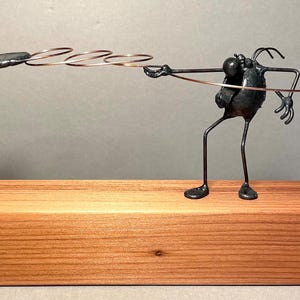 May include: A whimsical metal sculpture depicting a disc golf scene. A stylized figure made of dark metal wire throws a disc towards a miniature disc golf basket, also crafted from dark metal. The sculpture is mounted on a light brown wooden base. The artwork showcases intricate details and a playful design, suitable for disc golf enthusiasts or as a unique decorative piece.