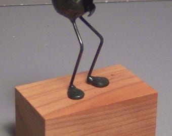 May include: A quirky metal sculpture of a creature with a rounded body, slender legs, and a curved antenna. The dark green figure stands on a rectangular wooden base, showcasing an artistic design.