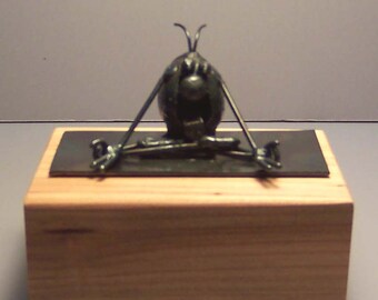 May include: A metal sculpture of a cartoon character sitting in a yoga pose on a wooden box with a black top.