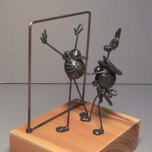 May include: A metal sculpture of two figures, one with arms raised, standing in front of a metal frame. The sculpture is mounted on a wooden base.