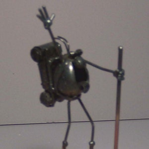 May include: A metal sculpture of a person with a backpack and a walking stick. The sculpture is standing on a wooden base.